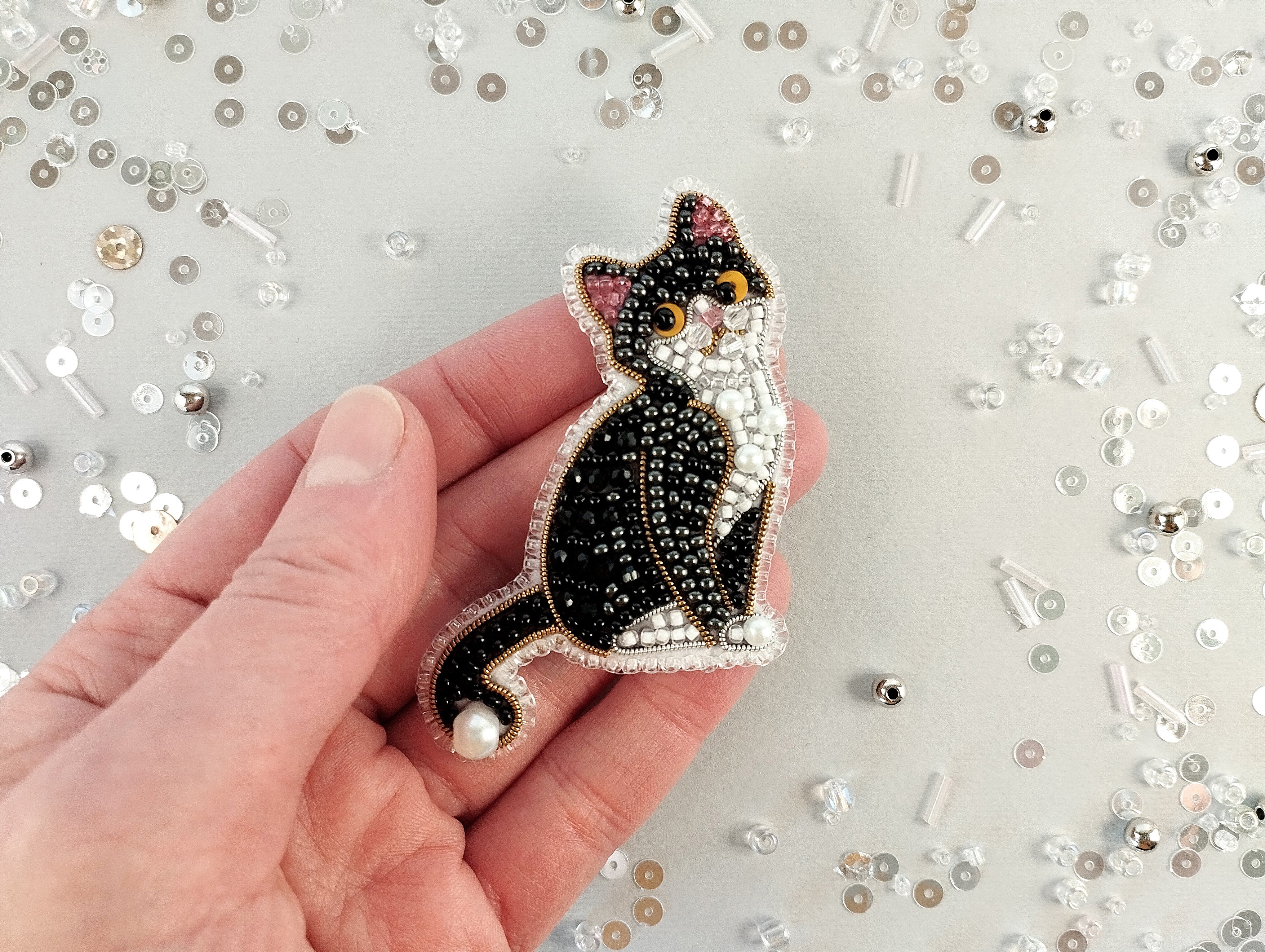 A hand holding a decorative beaded cat pin against a background of scattered beads and sequins.